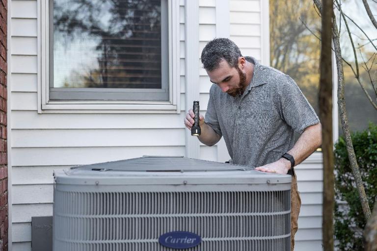 Technician inspecting an outdoor air conditioning unit with a flashlight next to a home window